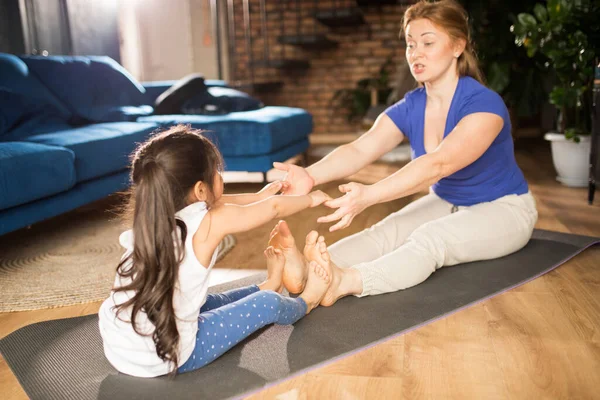 Girl reaching to her mother while stretching at the mat at the floor — Stock Photo, Image