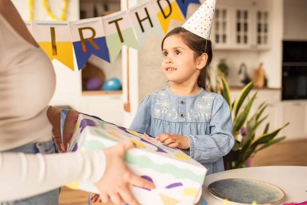 Chica usando sombrero de fiesta mirando a su madre mientras acepta su regalo de cumpleaños — Foto de Stock