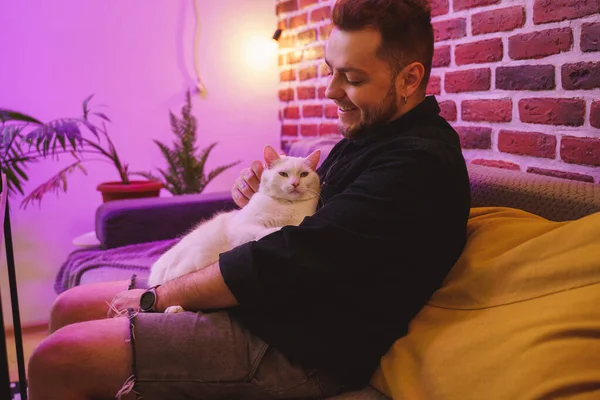 Man sitting at the sofa with his white cat at the laps and smiling happily — Stock fotografie