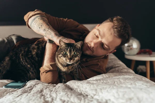 Hombre acariciando a su gato rayado mientras pasa tiempo con él en el dormitorio —  Fotos de Stock