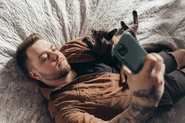 Egy férfi fekszik az ágyon a macskájával és szelfit csinál. — Stock Fotó