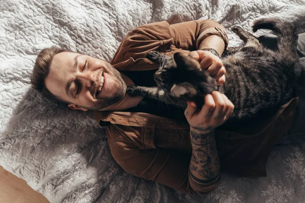 Man laughing and stroking his striped cat while spending time with him at bedroom — Stock fotografie