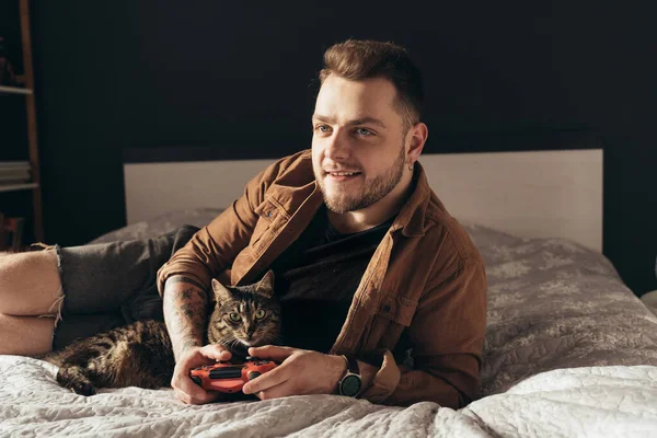 Man laying at the bed with his striped cat and playing at the video games — Φωτογραφία Αρχείου