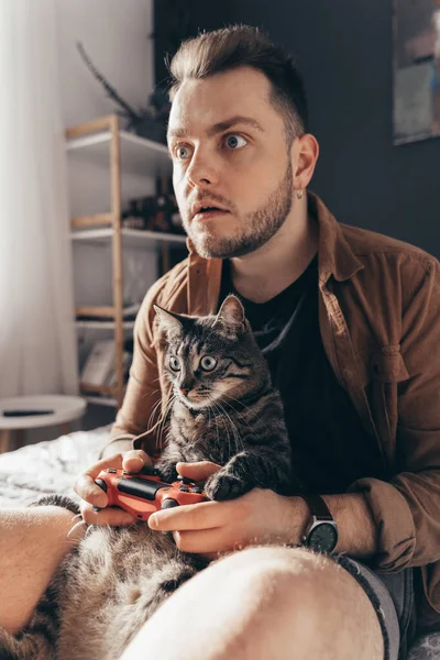 Man passionately playing at the video game while sitting at bed with his cat — Φωτογραφία Αρχείου