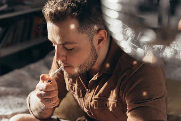 Hombre enciende un cigarrillo enrollado a mano con un encendedor mientras fuma cannabis en casa — Foto de Stock