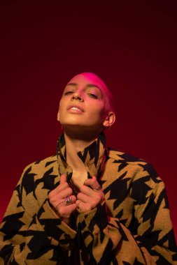 Şık bir ceket giyen bir kadın stüdyoda dikilirken yakasını düzeltiyor.