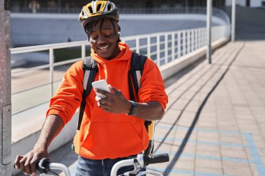 Kasklı ve teslimat çantalı bir adam akıllı telefon ekranına bakıyor.