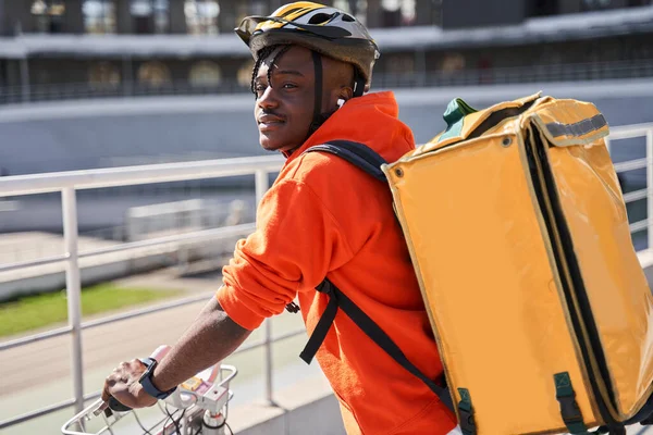 คนส่งของใส่เสื้อสเวตเตอร์สีแดงและกระเป๋าเป้สะพายหลังสีเหลือง นั่งที่จักรยาน — ภาพถ่ายสต็อก
