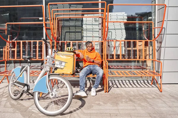 Dostawca żywności siedzi na stacji przy ulicy w pobliżu swojego plecaka rowerowego i termo — Zdjęcie stockowe