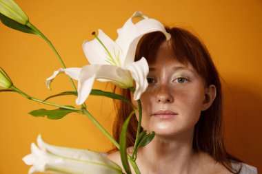 Stüdyoda poz verirken yüzünü çiçeklerin arkasına saklayan kızıl kadın.