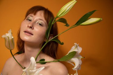 Kızıl model, çıplak omuzlu, zambak tutan ve kameraya bakan.