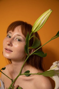 Çıplak makyajlı bir kadın zambak buketi tutuyor ve kameraya bakıyor.