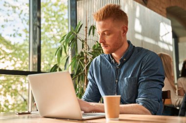 Kotlu tişört giyen bir adam kafede oturup çalışırken odaklanıyor.