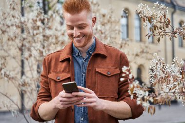 Çiçek açan ağaçların etrafında duran ve akıllı telefonuna bakan bir adam.