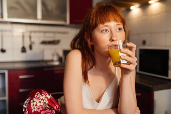 Mujer bebiendo jugo de naranja y pensando en algo mientras mira a la ventana — Foto de Stock