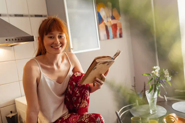 Frau liest Buch, während sie in der gemütlichen Küche sitzt — Stockfoto