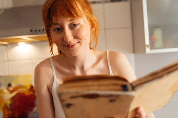 Glad ingefära kvinna läser bok, medan du sitter i det mysiga köket — Stockfoto