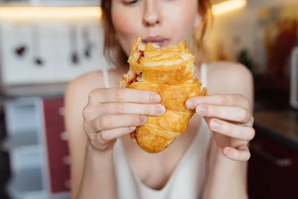 Ingwermädchen isst Croissant mit Appetit beim Frühstück in der hellen Küche — Stockfoto