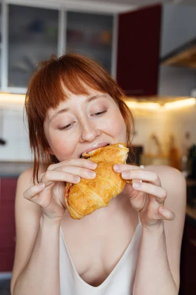 Girl eating croissant with appetit while having breakfast at the light kitchen — Stock Photo, Image