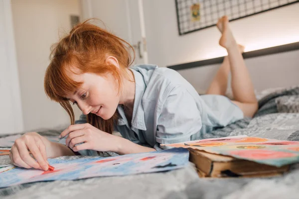 年轻女人坐在床边，在公寓里画画 — 图库照片