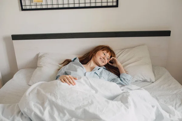 Chica pelirroja tendida debajo de la manta y despertando por la mañana en casa —  Fotos de Stock