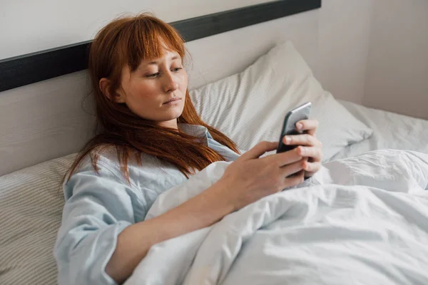 Žena ležící pod dekou a prohlížení internetu na svém smartphonu — Stock fotografie