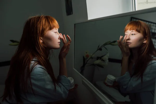 女人一边擦着她的皮肤，一边用润肤霜，同时看着镜子 — 图库照片