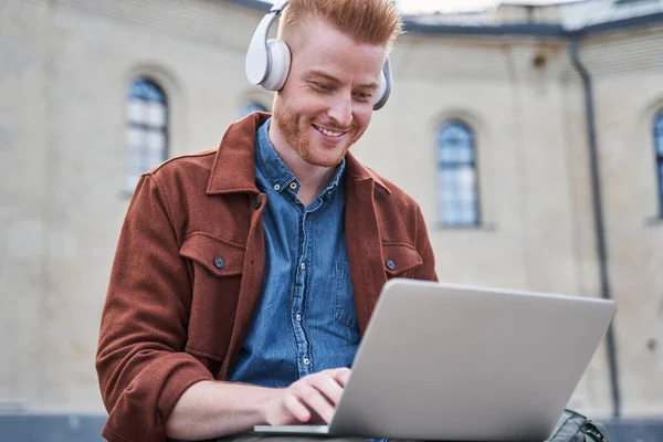 Mann mit Kopfhörer sitzt auf der Straße und tippt auf die Laptop-Tastatur — Stockfoto