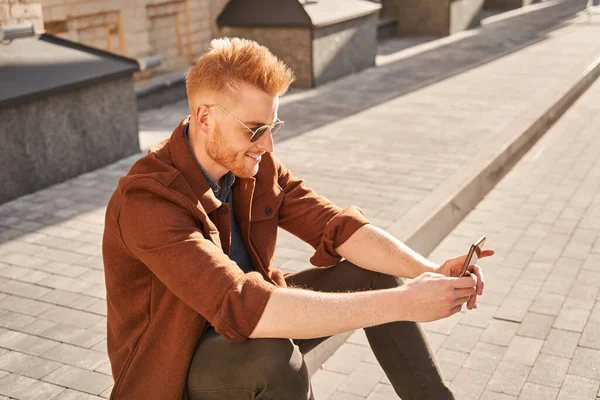 Чоловік в сонцезахисних окулярах сидить на сонці і дивиться на свій смартфон — стокове фото