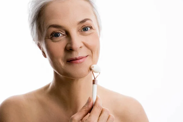 Mujer mirando a la cámara y sonriendo mientras masajea su mejilla con un rodillo de jade — Foto de Stock