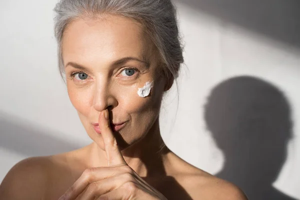 Kalme vrouw met hydraterende crème op haar kuiken met een vinger naar haar lippen — Stockfoto