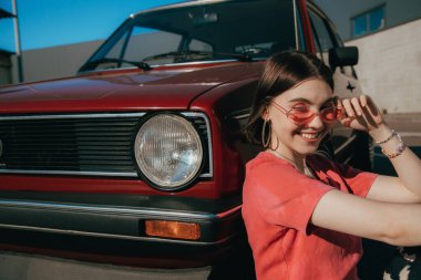 Pembe elbiseli kadın kırmızı arabanın yanında oturuyor ve şık poz veriyor.