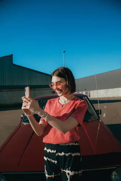 Woman wearing sunglasses holding smartphone and making selfie near her car — Stock Photo, Image