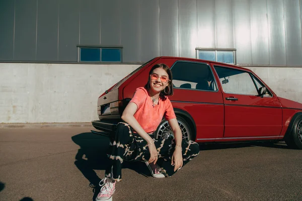 Mädchen mit Brille blickt in die Kamera und lächelt, während sie neben dem Auto sitzt — Stockfoto