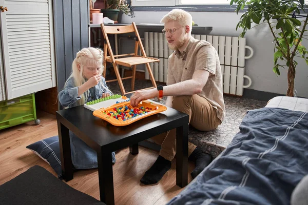 Menina dobrando detalhes de seu jogo e sentado à mesa, enquanto seu pai loiro ajudando — Fotografia de Stock