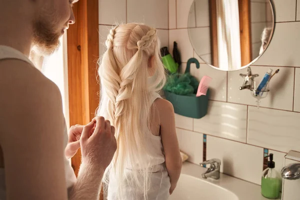 Mężczyzna czesający włosy swojej małej blondynki w łazience i tkający warkocze — Zdjęcie stockowe