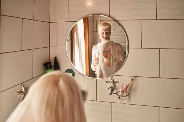 Man doing hairstyle for his little daughter at the bathroom while smiling