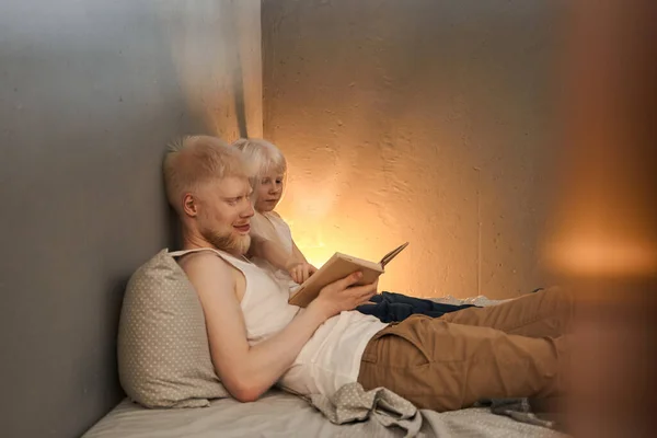 Man laying at the bed with his little blonde daughter and reading book for her — ストック写真