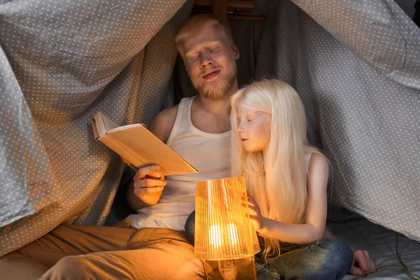 男座っている彼の小さなアルビノの娘と読書本のランプの光によって — ストック写真