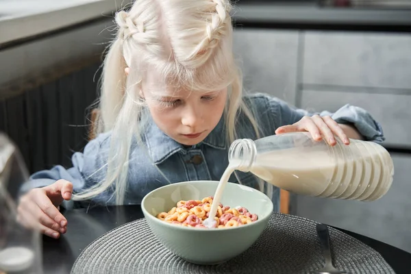 Κορίτσι ρίχνει γάλα από το μπουκάλι στο πιάτο, ενώ κάθεται στην κουζίνα — Φωτογραφία Αρχείου