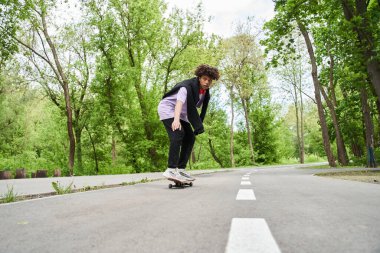 Kıvırcık saçlı bir adam yaz sokağında kaykayla şarkı söylüyor.