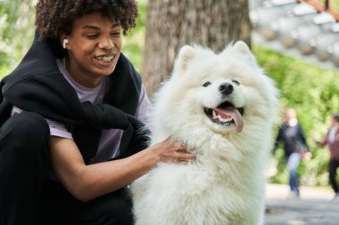 Adam dişlek gülümsüyor, sokakta dizlerinin üzerinde oturuyor ve beyaz tüylü köpeğini okşuyor.