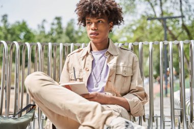 Çocuk parkta bankta oturuyor ve deftere bir şeyler yazıyor.