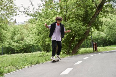 Kıvırcık saçlı bir adam yaz sokağında kaykayla şarkı söylüyor.