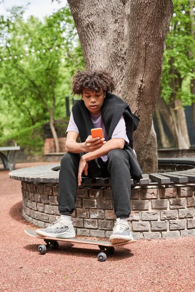 Ragazzo seduto alla panchina e tenendo le gambe allo skateboard durante la navigazione internet — Foto Stock