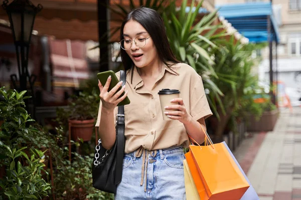 Mujer leyendo un mensaje conmocionado mientras bebía café y de pie con sus compras — Foto de Stock