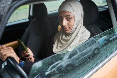 Kadın arabaya biniyor ve akıllı telefonunu kullanırken pencereye bakıyor.