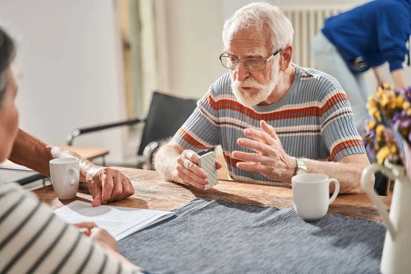 Senior man die aan de kaarten speelt met zijn vrienden terwijl hij aan tafel zit — Stockfoto