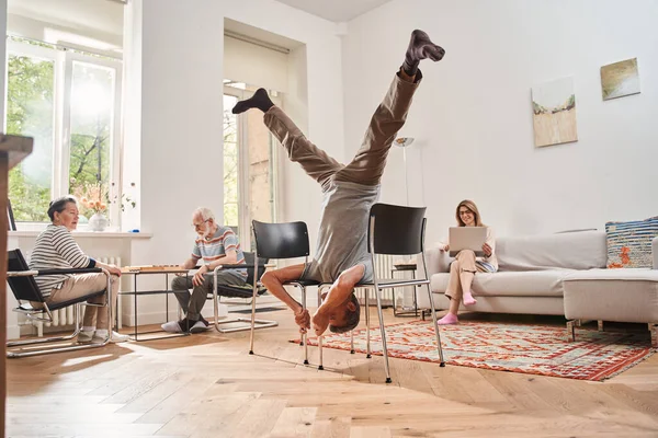 Starší muž stojí hlavou dolů na židlích, zatímco dělá akrobatické kousky — Stock fotografie