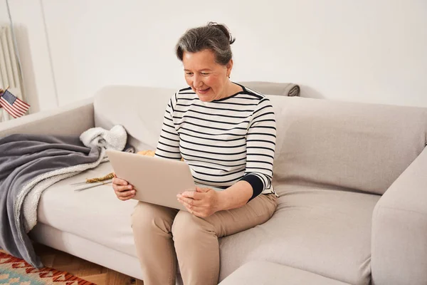 Старша жінка тримає ноутбук на колінах і посміхається під час спілкування з родичами — стокове фото